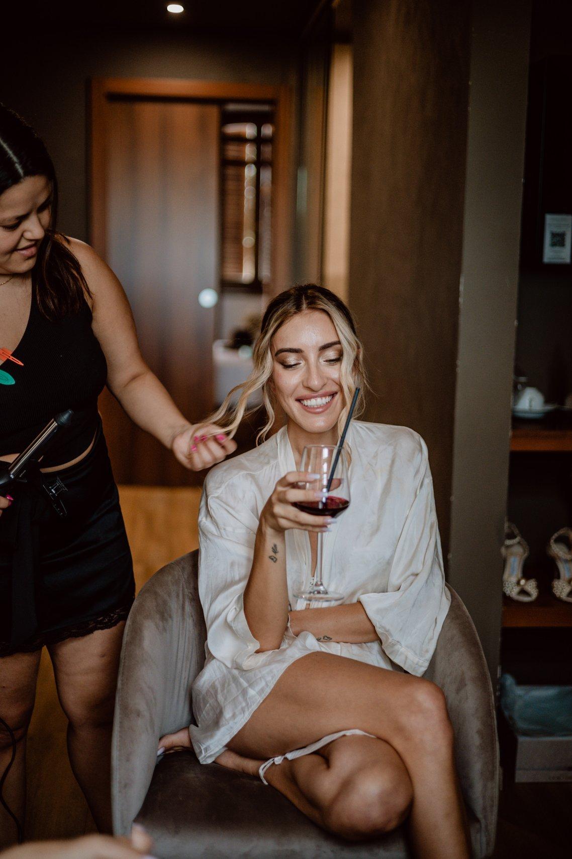 sposa con un bicchiere di vino in mano che viene pettinata da una hairstylist